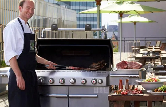 events-bbq-in-side Koch steht auf der Terrasse unter einem grünen Sonnenschirm und bereitet Fleisch auf einem Grill zu