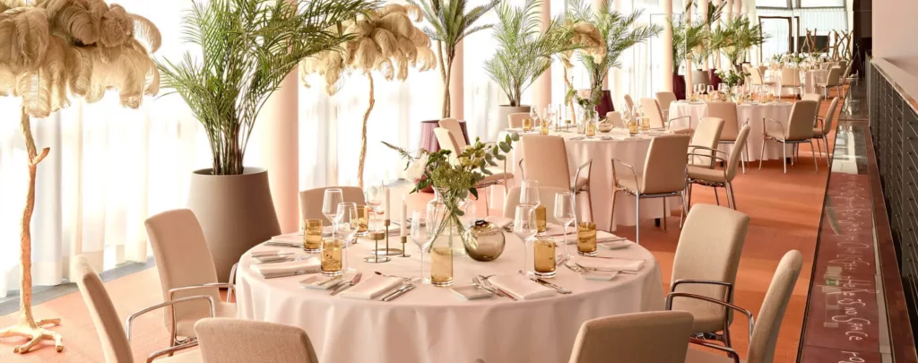 Elegantes Ambiente in einem Restaurant und Bar in Hamburg mit runden, weiß gedeckten Tischen, Palmen und einer Federdeko.